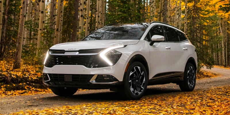 An image of a white Kia Sportage driving through a forested area in the autumn, surrounded by yellow leaves, with its daytime running lights on. 