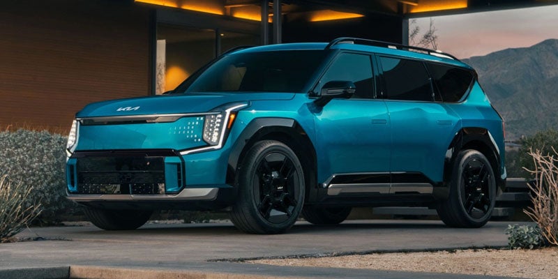 A metallic blue Kia EV9 sitting on a parking pad in front of a home with a painting of a mountain vista behind it. 