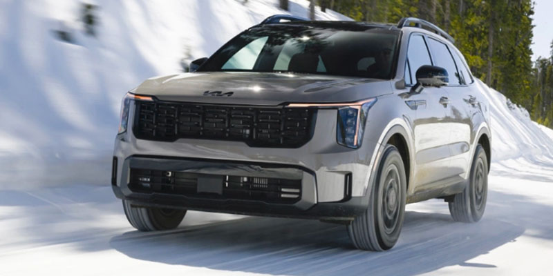 A silver Kia Sorento driving through a snowy landscape with evergreen trees in the background.