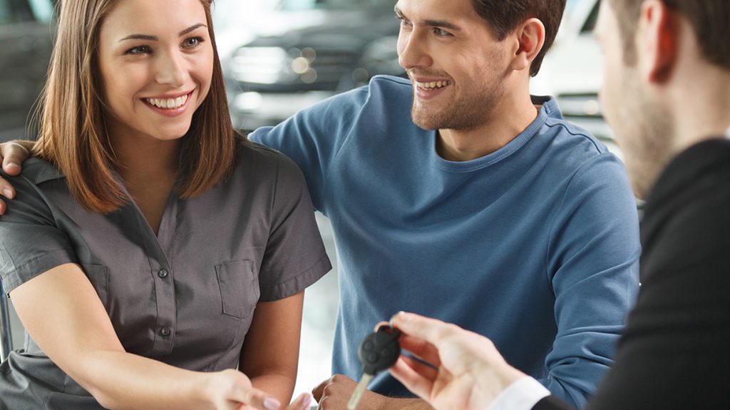 An image of a couple getting the key to their new vehicle after completing the dealership financing process. They are both smiling and happy to be driving their new car home. 