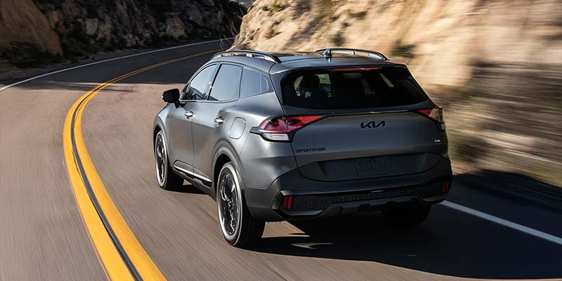 A grey 2026 Kia Sportage Hybrid exterior as seen from the rear and driver's side drives down a highway near Shiloh, IL