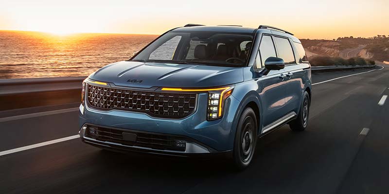 a blue 2026 Kia Carnival minivan drives down a road next to a body of water near Shiloh, IL