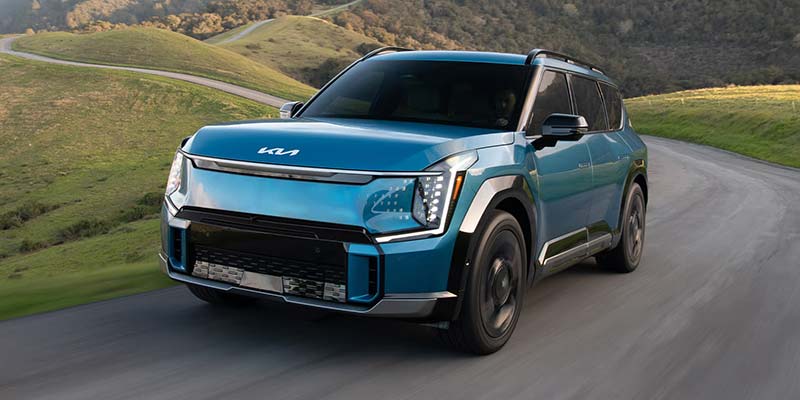 Blue Kia EV9 driving on a winding coastal road with a dramatic ocean and cliffside background.